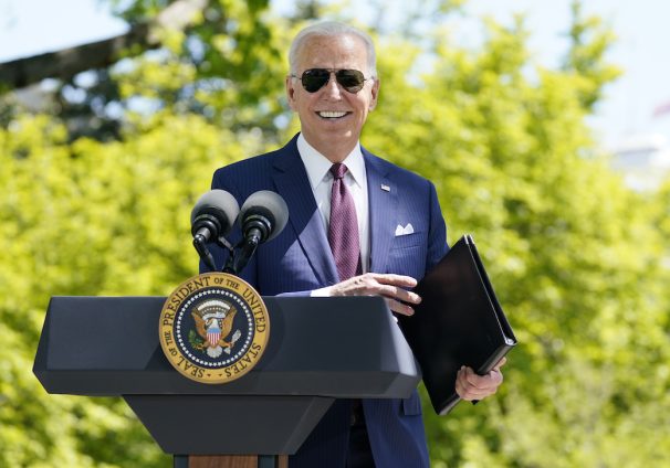 President Joe Biden, smiling with aviator sunglasses, holds a folder while standing behind a lectern. Green trees are in the background.