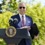 President Joe Biden, smiling with aviator sunglasses, holds a folder while standing behind a lectern. Green trees are in the background.